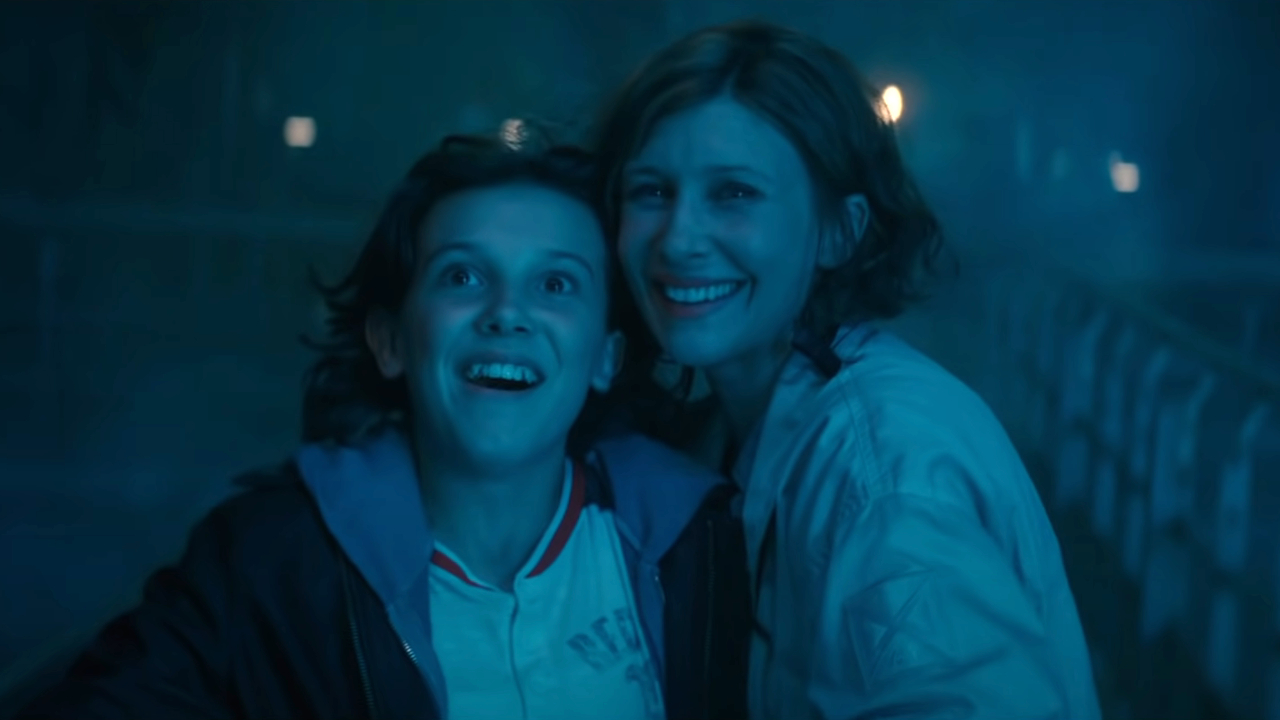 Millie Bobbie Brown und Vera Farmiga stehen lächelnd im blauen Licht in Godzilla: King of the Monsters.