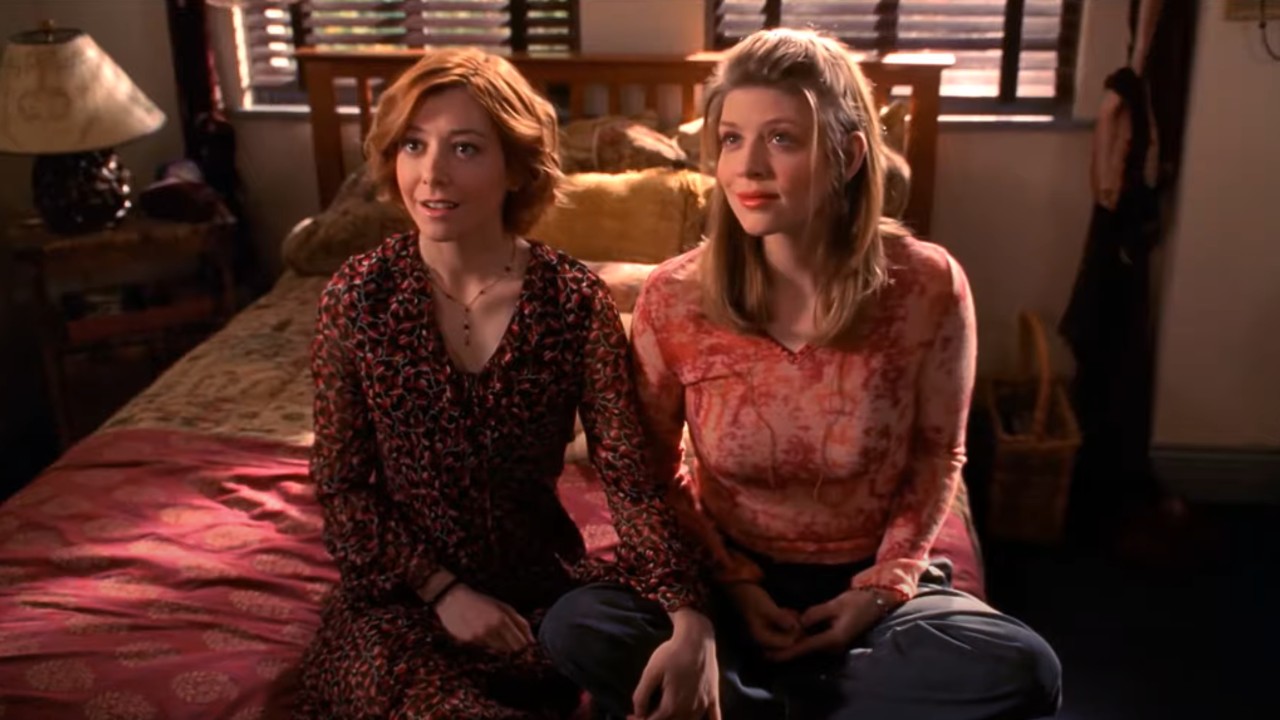 Willow und Tara halten Händchen, während sie auf einem Bett in „Buffy – Im Bann der Dämonen“ sitzen.